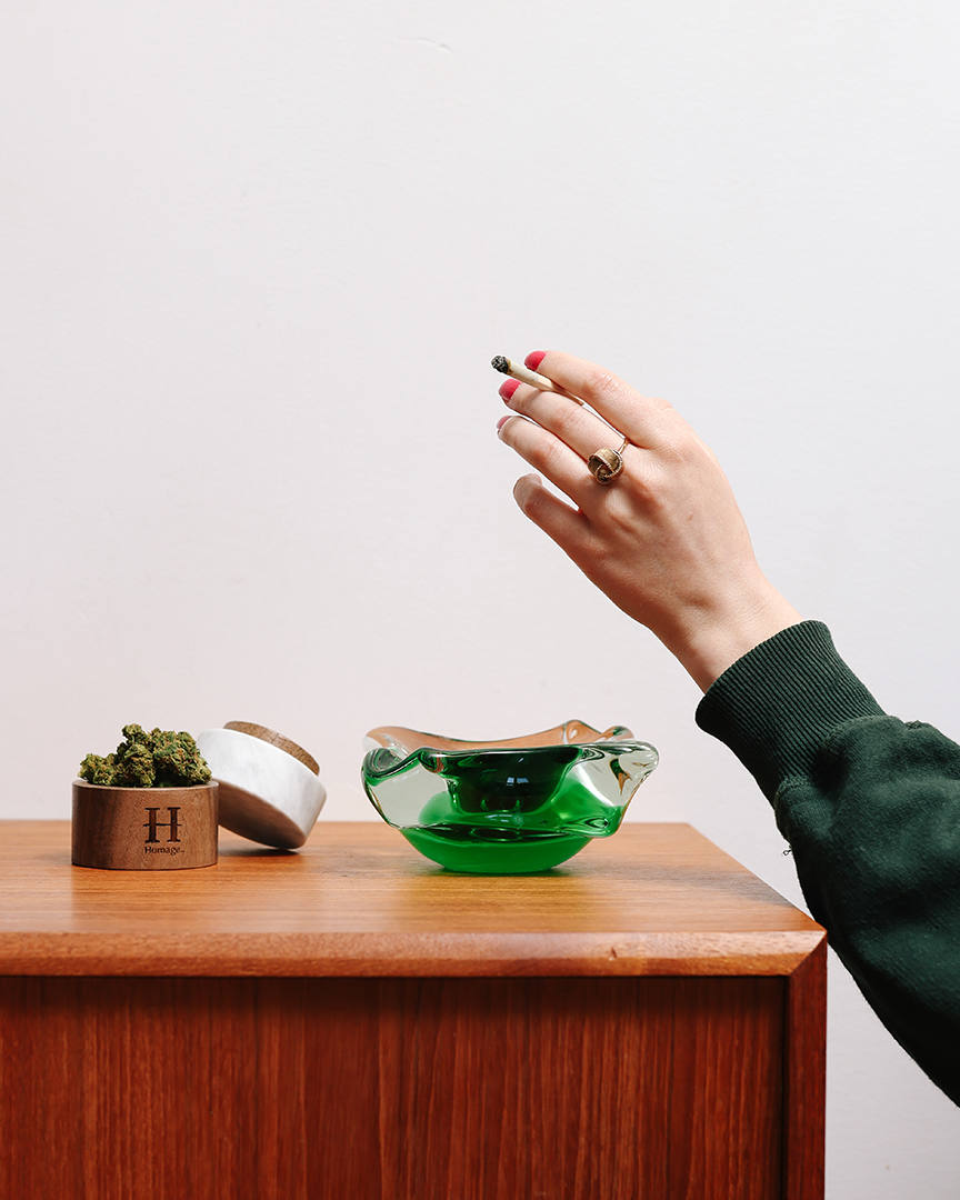 main de femme élégante tenant un joint appuyée contre un meuble sur lequel se trouvent un cendrier et un contenant de cannabis Homage et quelques fleurs séchées